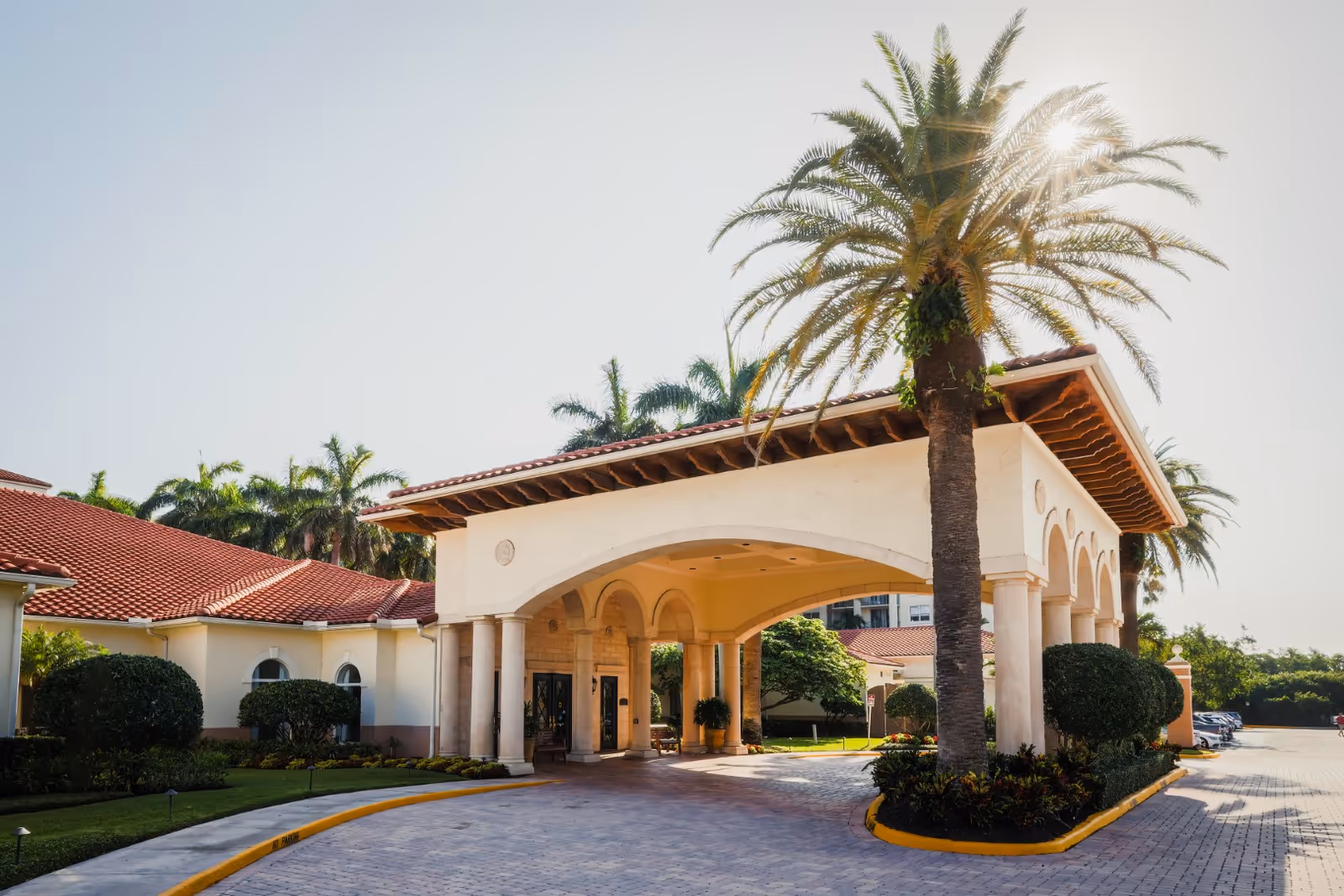 Entrance area of a senior living facility with a covered driveway supported by columns, surrounded by palm trees and well-maintained landscaping under a clear sky.