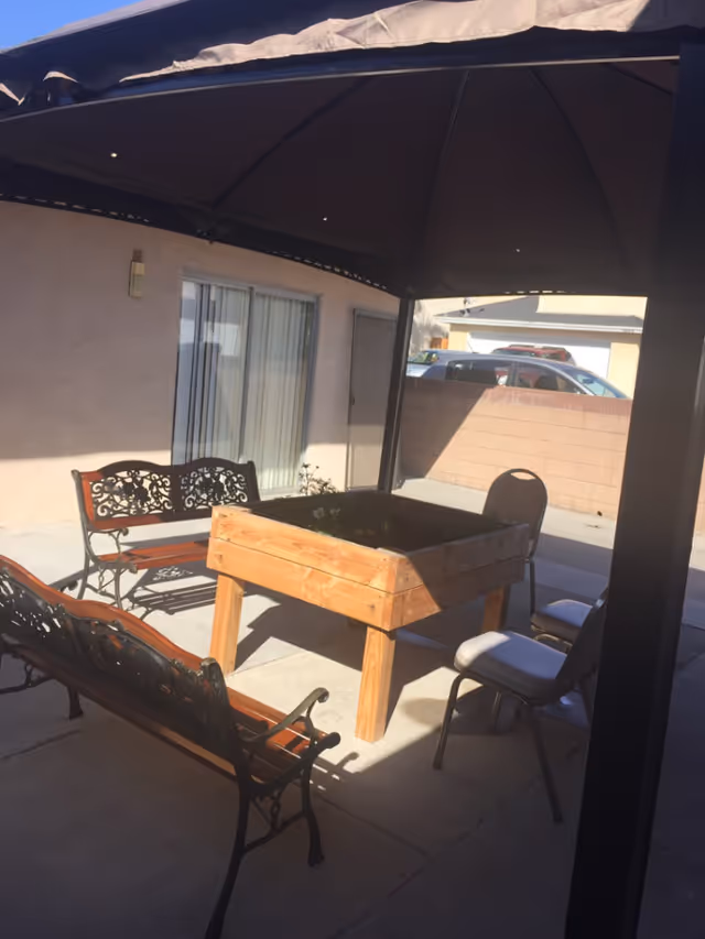 Outdoor patio area with a wooden raised garden bed in the center, surrounded by two metal and wood benches and two cushioned chairs. The area is shaded by a large canopy. In the background, there is a beige building with a sliding glass door and a window, and a brick wall with parked cars visible beyond it.