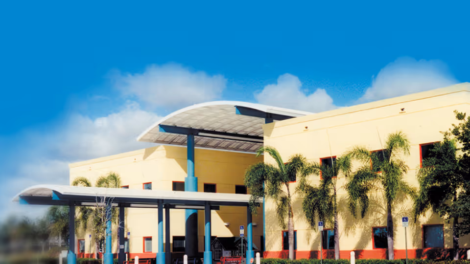 Exterior view of a yellow building with blue columns, palm trees, and a covered entrance.
