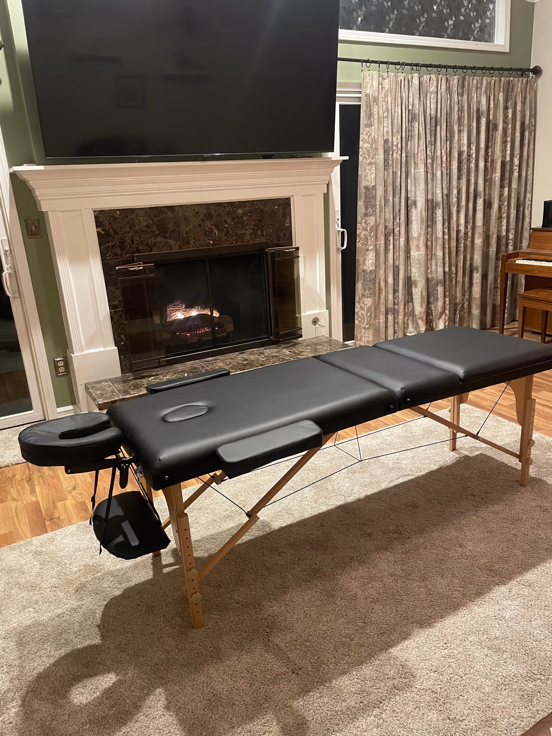 A black massage table setup in a living room with fireplace, curtains, and a mounted TV.