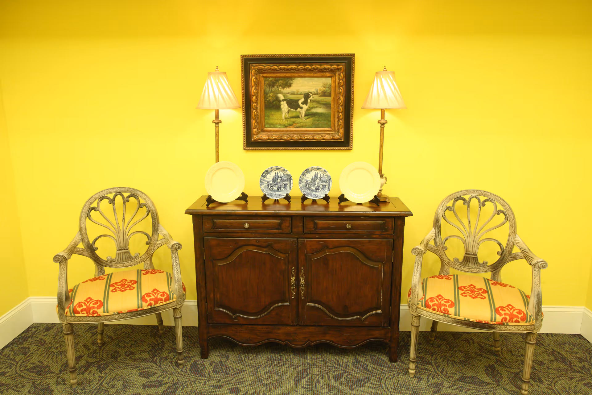 Two decorative chairs flank a dark wooden sideboard topped with plates, lamps, and a framed painting against a yellow wall.