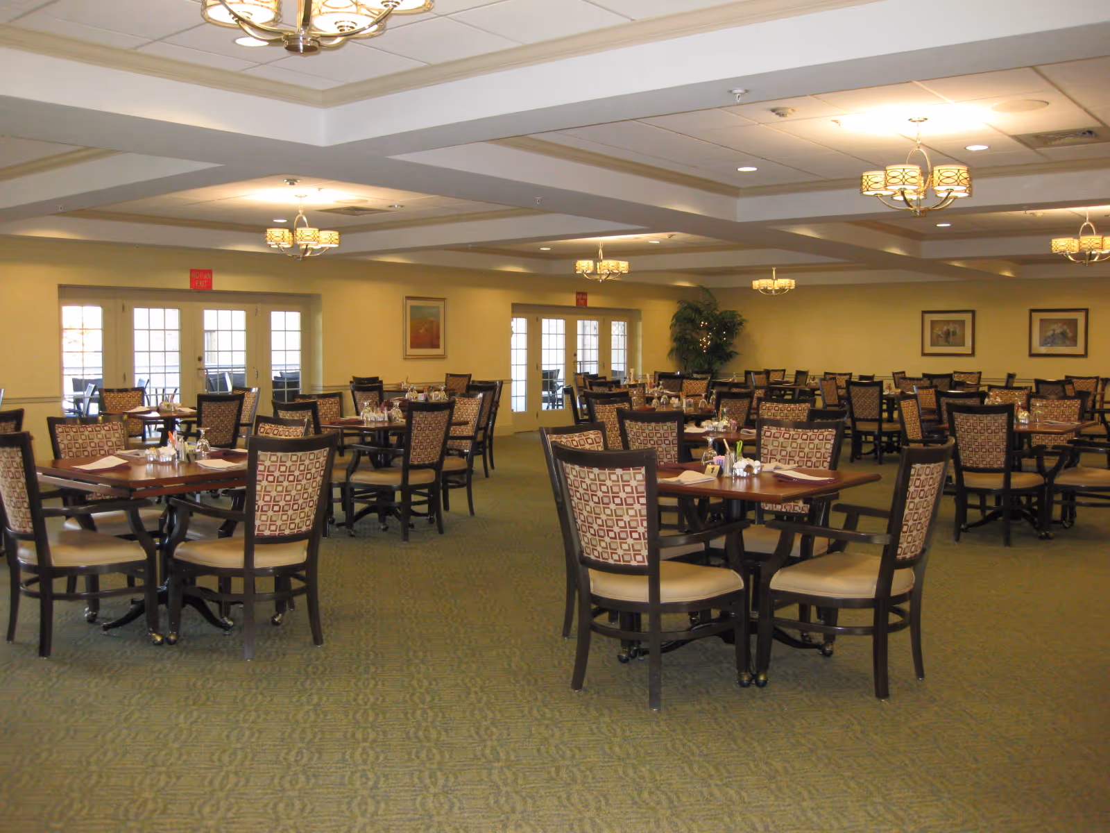 A spacious dining room with multiple square wooden tables and cushioned chairs arranged neatly. The room has green carpet flooring, beige walls adorned with framed artwork, and several ceiling light fixtures. French doors lead to an outdoor patio area.