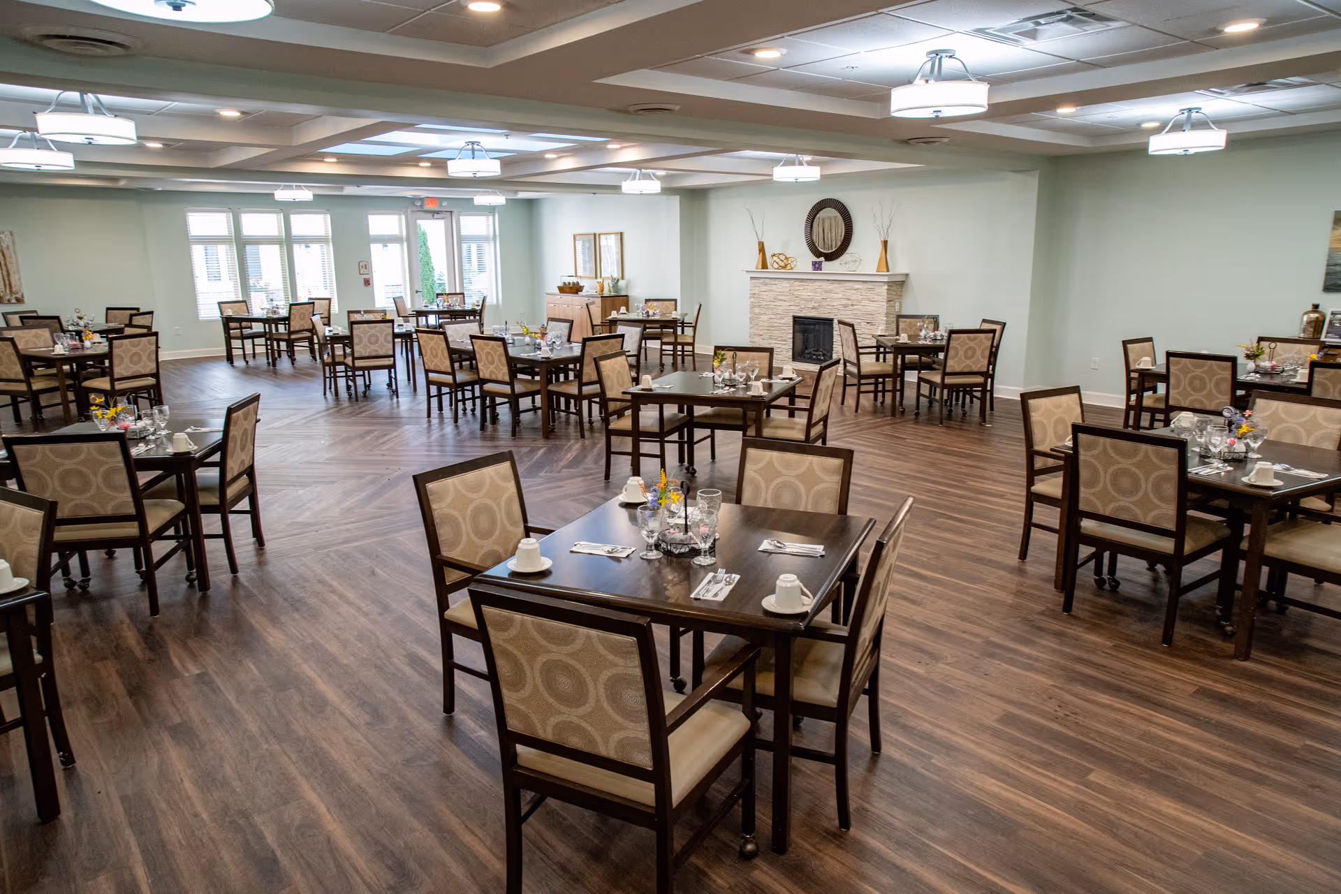A spacious dining room with multiple dark wood tables and cushioned chairs arranged neatly. Each table is set with cups, glasses, utensils, and small flower arrangements. The room has wood flooring, light-colored walls, ceiling lights, and a fireplace with decorative items on the mantel.