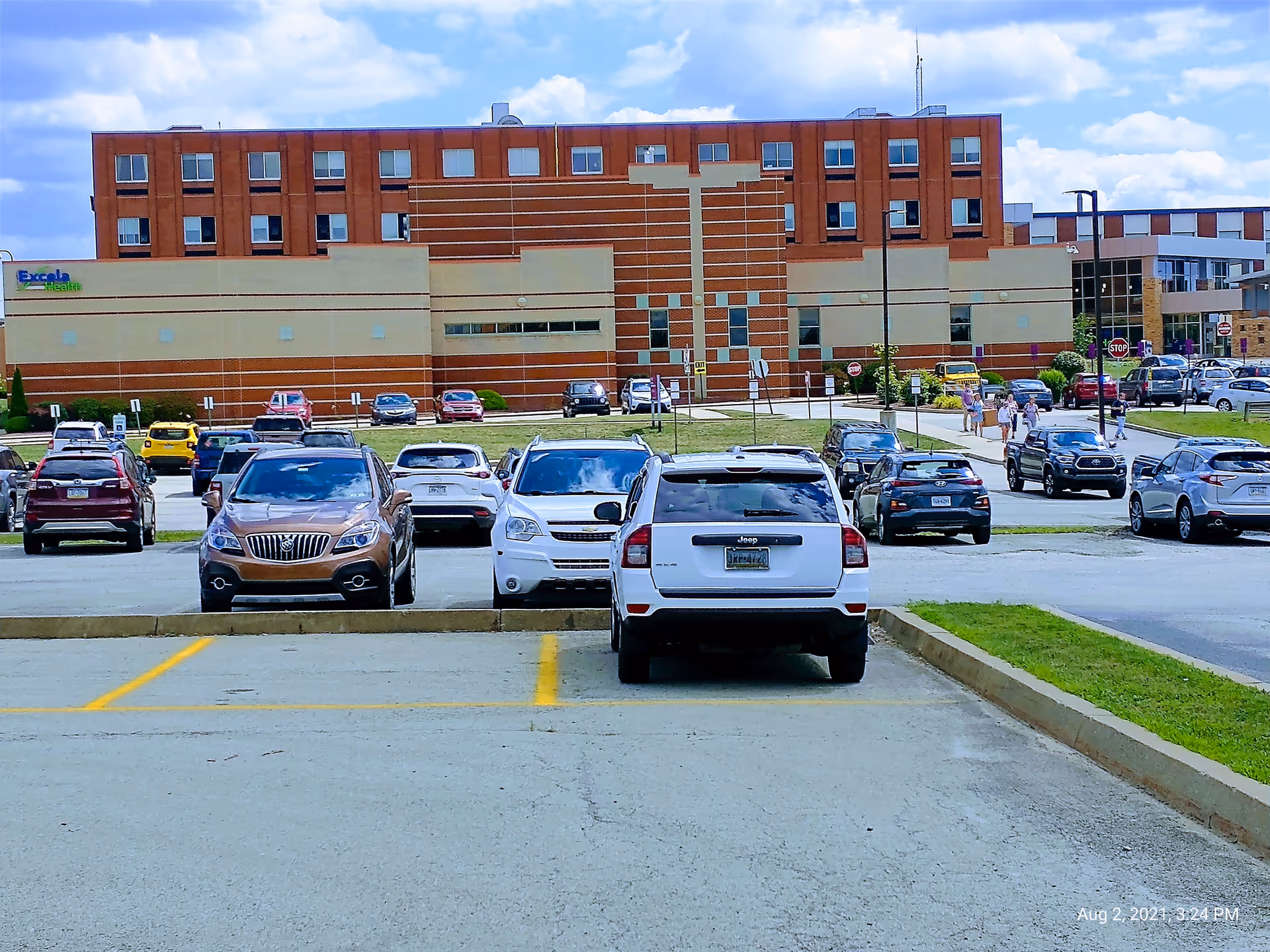 Front view of a brick hospital with a parking lot and many cars.