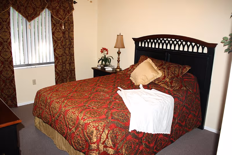 A bedroom with a large bed covered in a red and gold patterned bedspread, two matching pillows, and a beige pillow. A white garment is laid out on the bed. There is a wooden nightstand with a lamp, a small plant with red flowers, and some books. A window with vertical blinds and red patterned curtains is on the left wall.