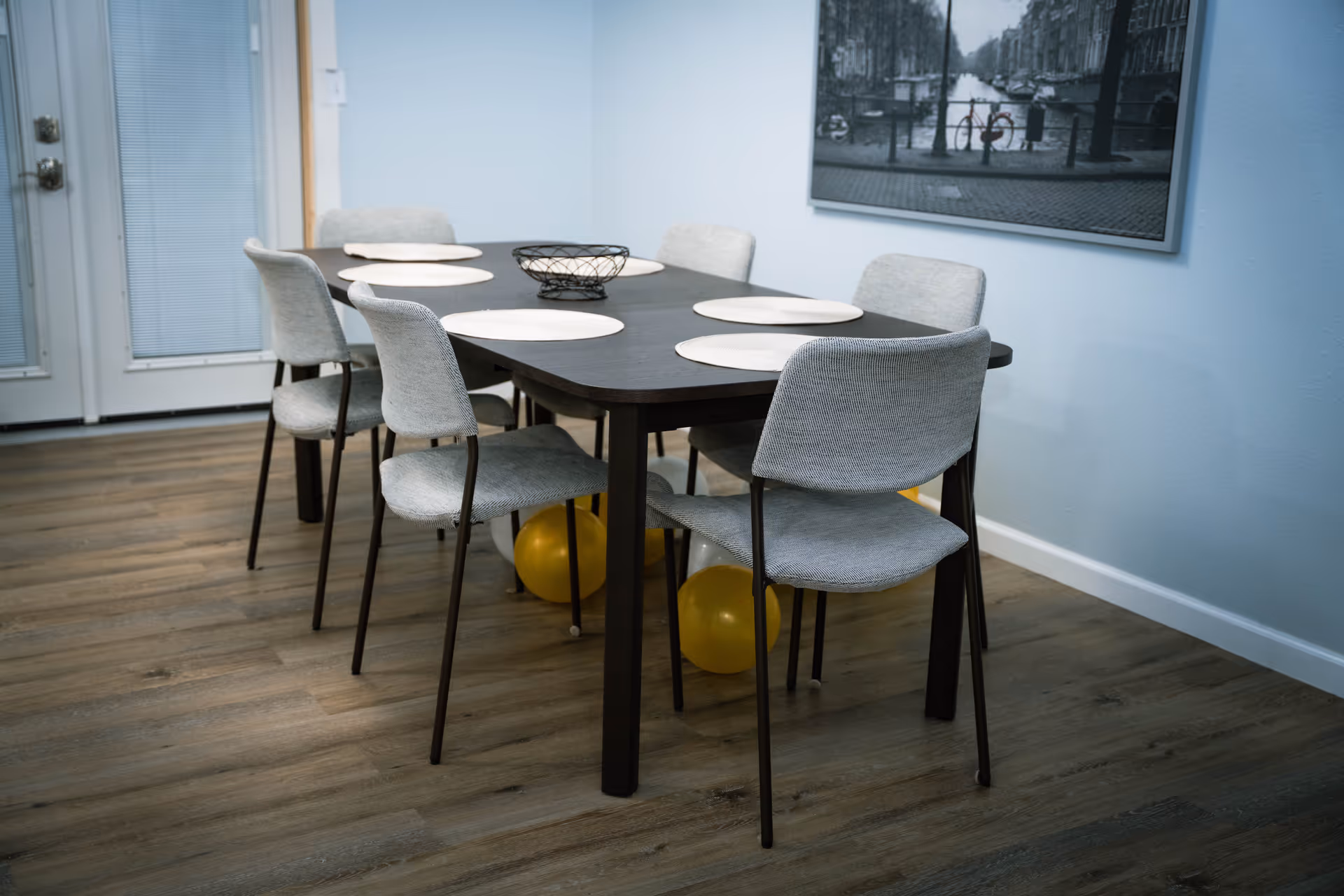 Dining room with a dark rectangular table, six gray upholstered chairs, round placemats and a decorative bowl on wood floors.