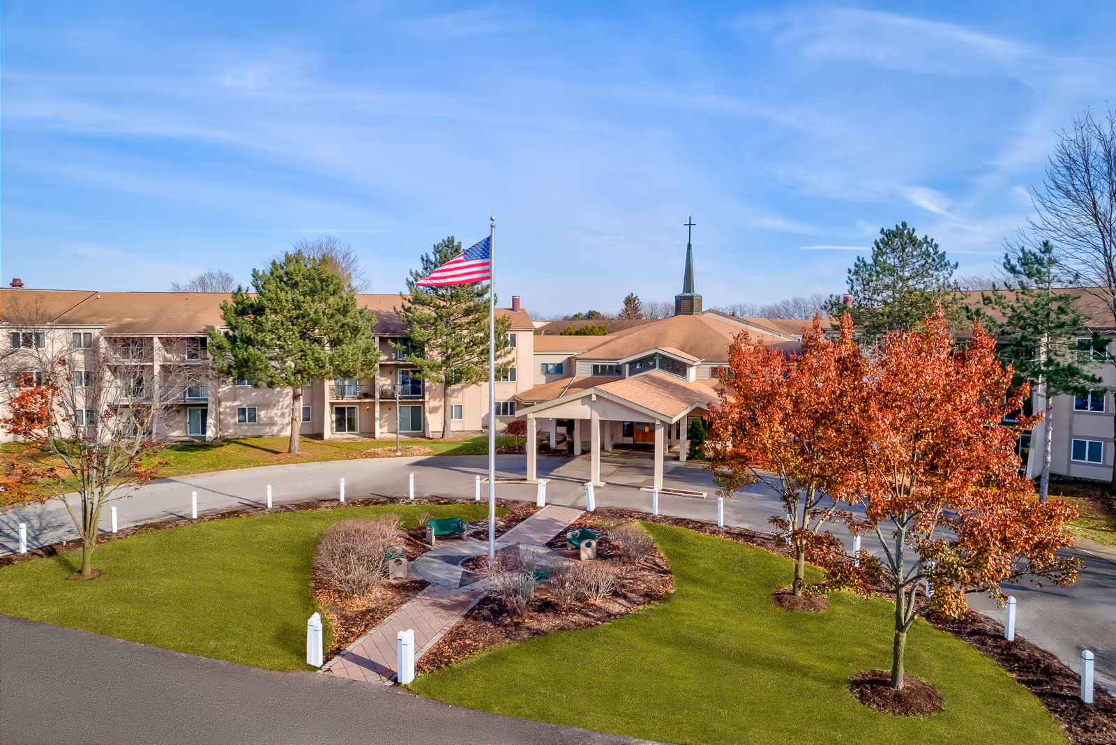 Front entrance of a multi-story senior living facility with a flagpole, circular driveway, and landscaped grounds.