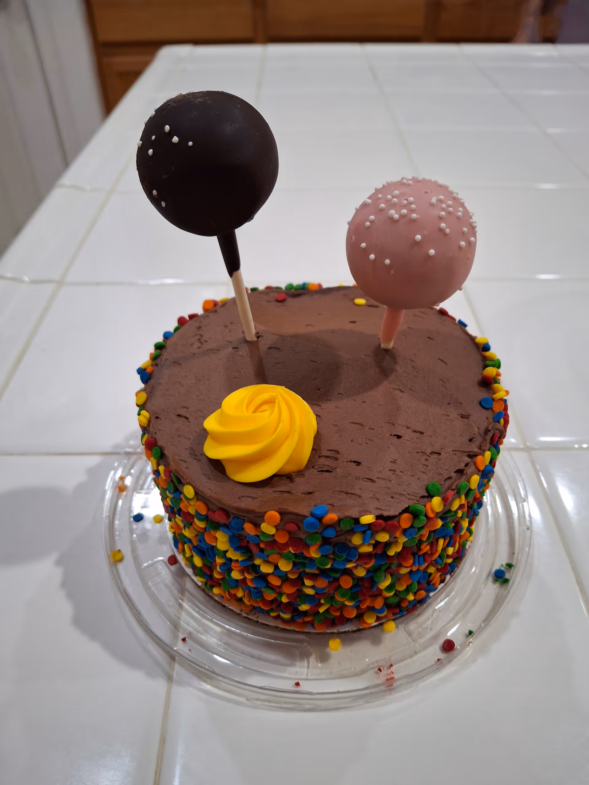 Chocolate cake with colorful sprinkles and two cake pops on a tiled kitchen counter