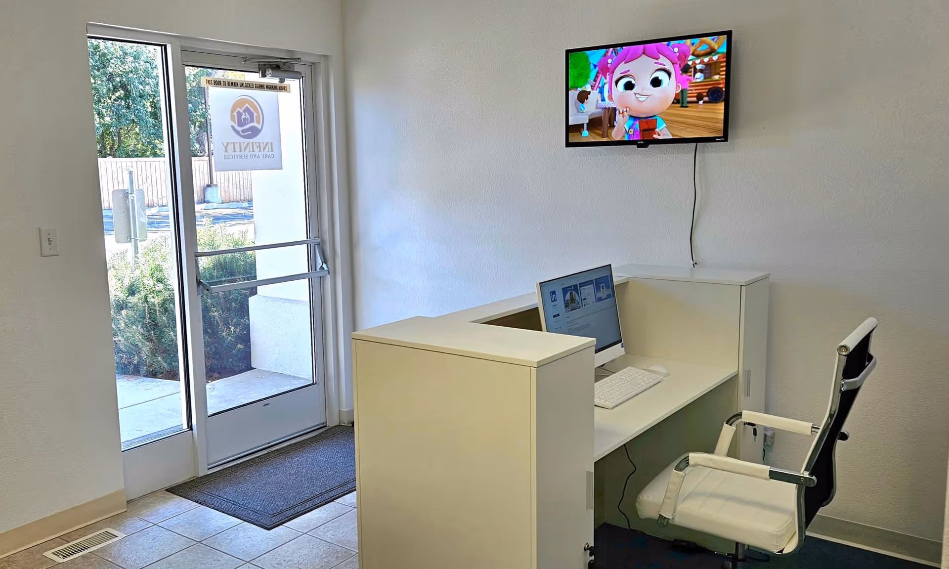 Interior reception area with white desk, computer, and wall-mounted TV near glass door.