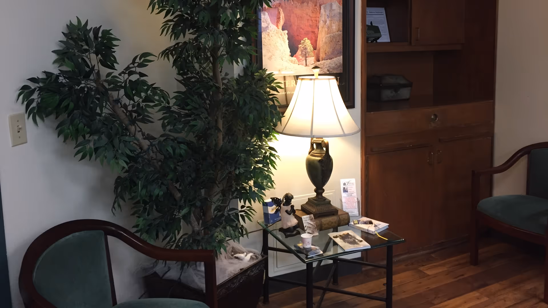 Lobby seating area with a glass table, lamp, large plant, and wood cabinet.