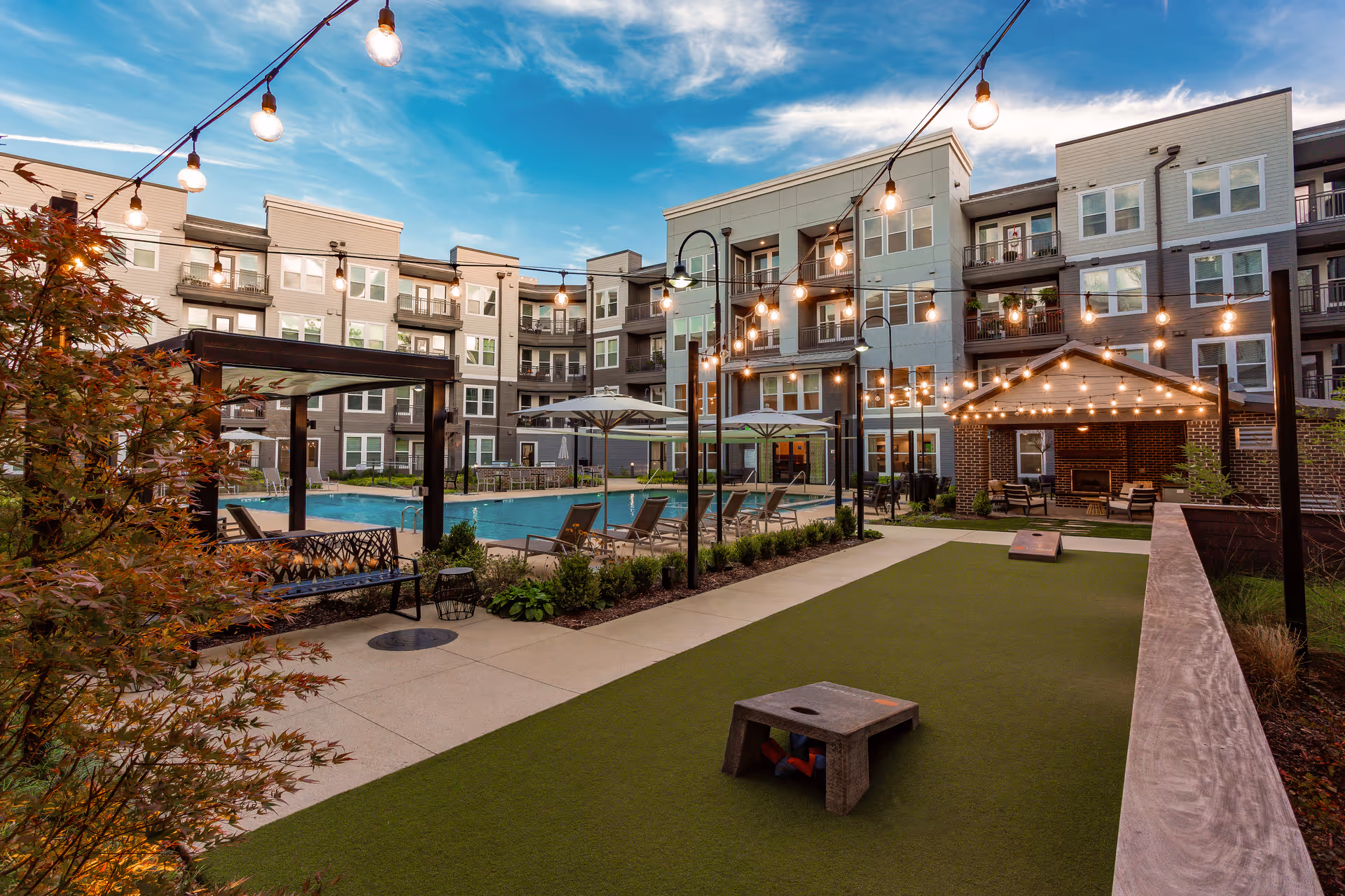 Outdoor courtyard area of a senior living facility with string lights hanging overhead, a swimming pool surrounded by lounge chairs and umbrellas, a covered seating area with a fireplace, and a bocce ball court in the foreground. The multi-story building with balconies surrounds the courtyard under a partly cloudy sky.