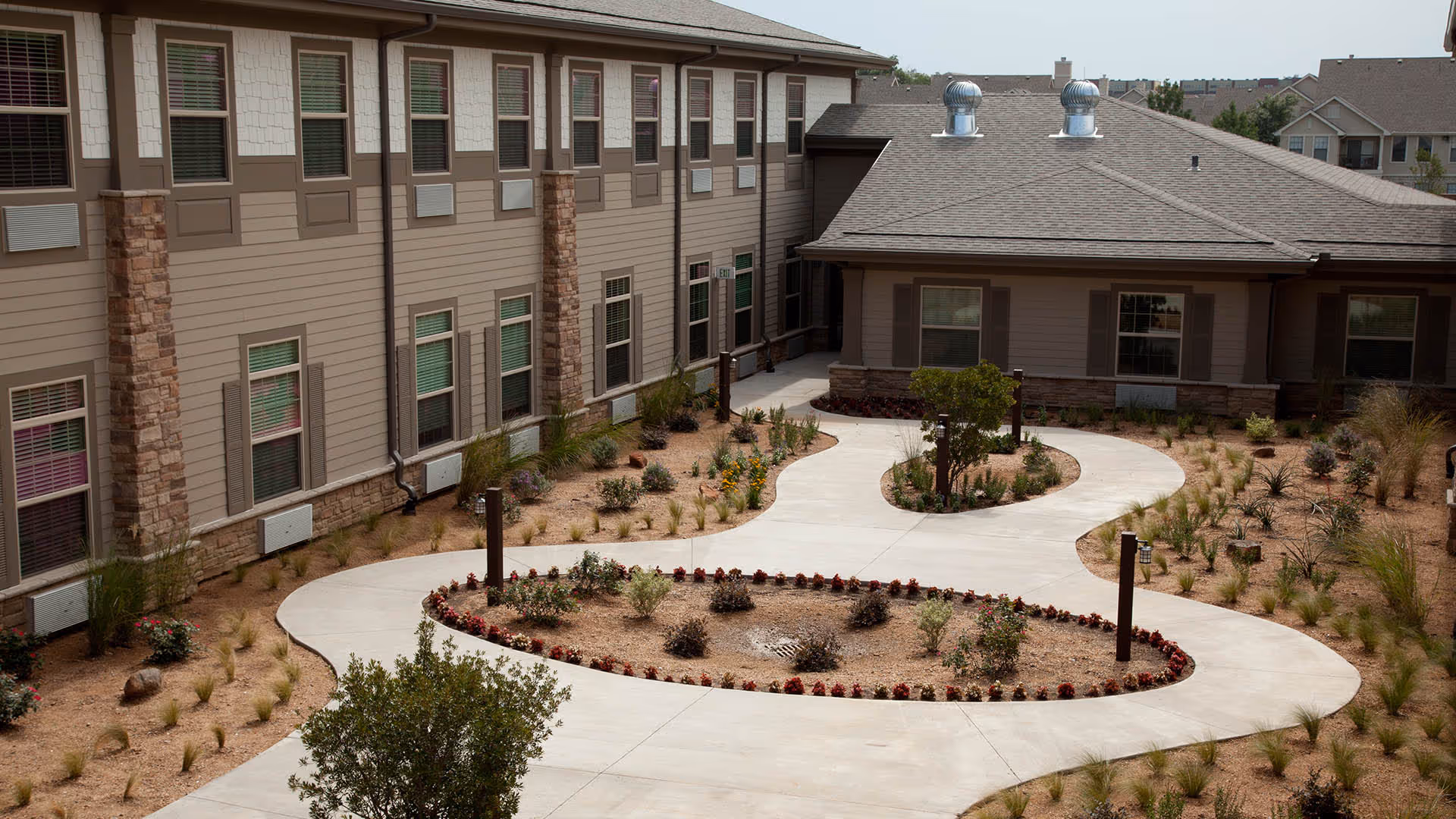 Curved concrete walkway winds through a landscaped courtyard beside a tan senior living building.