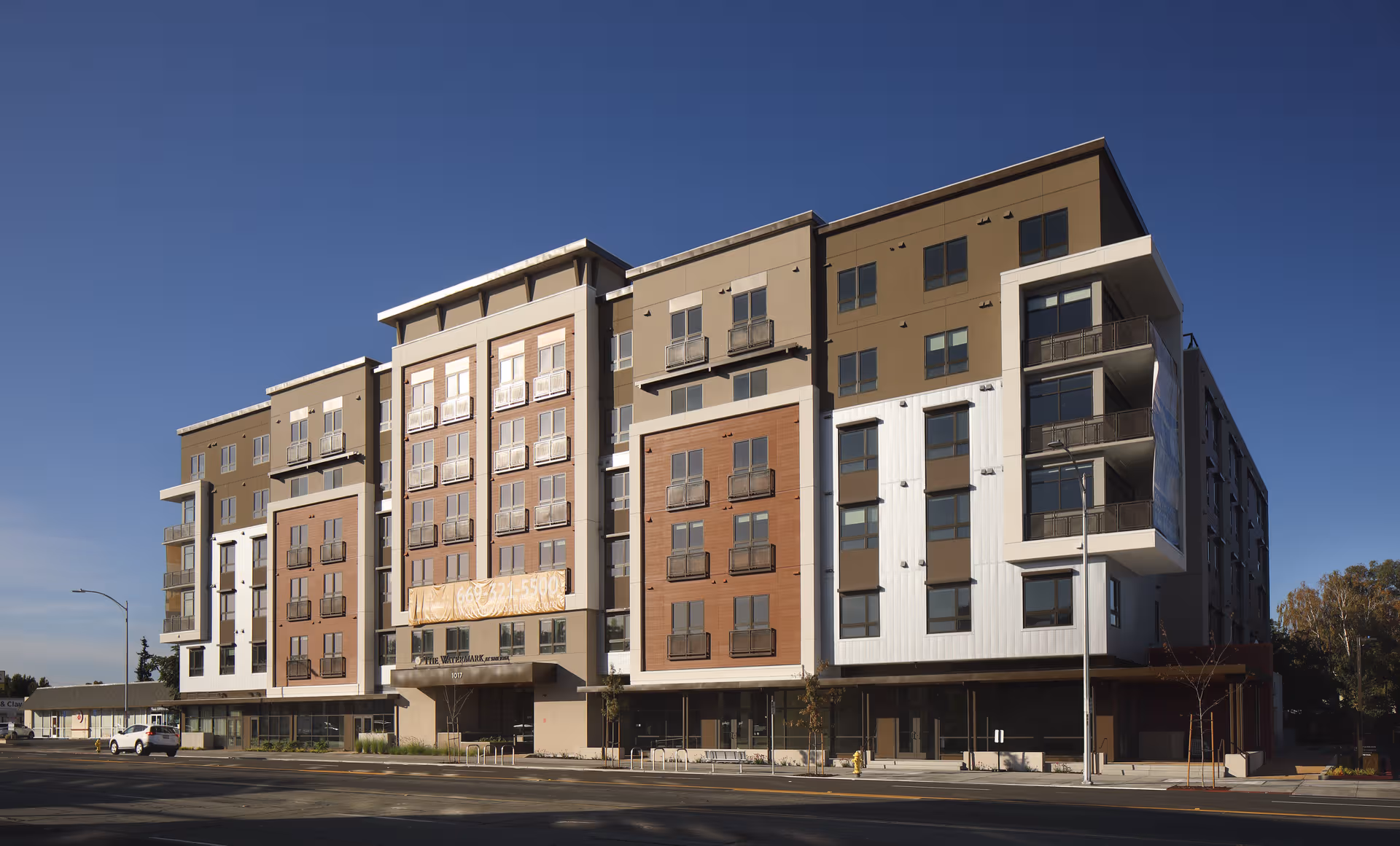 Modern senior living building with balconies along a wide street front under a clear blue sky.