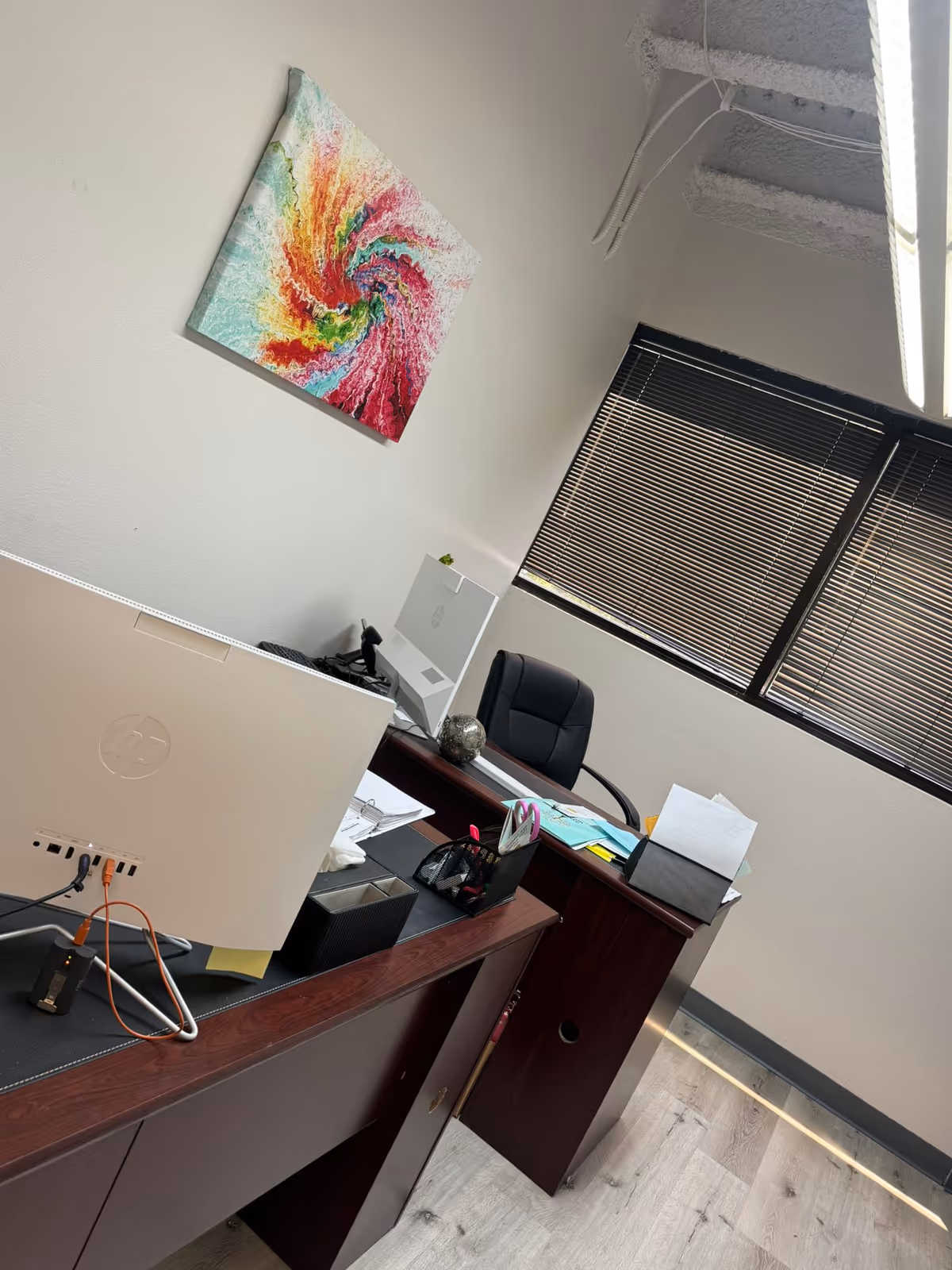 Office workspace with desk, computer, blinds, and colorful abstract wall art.