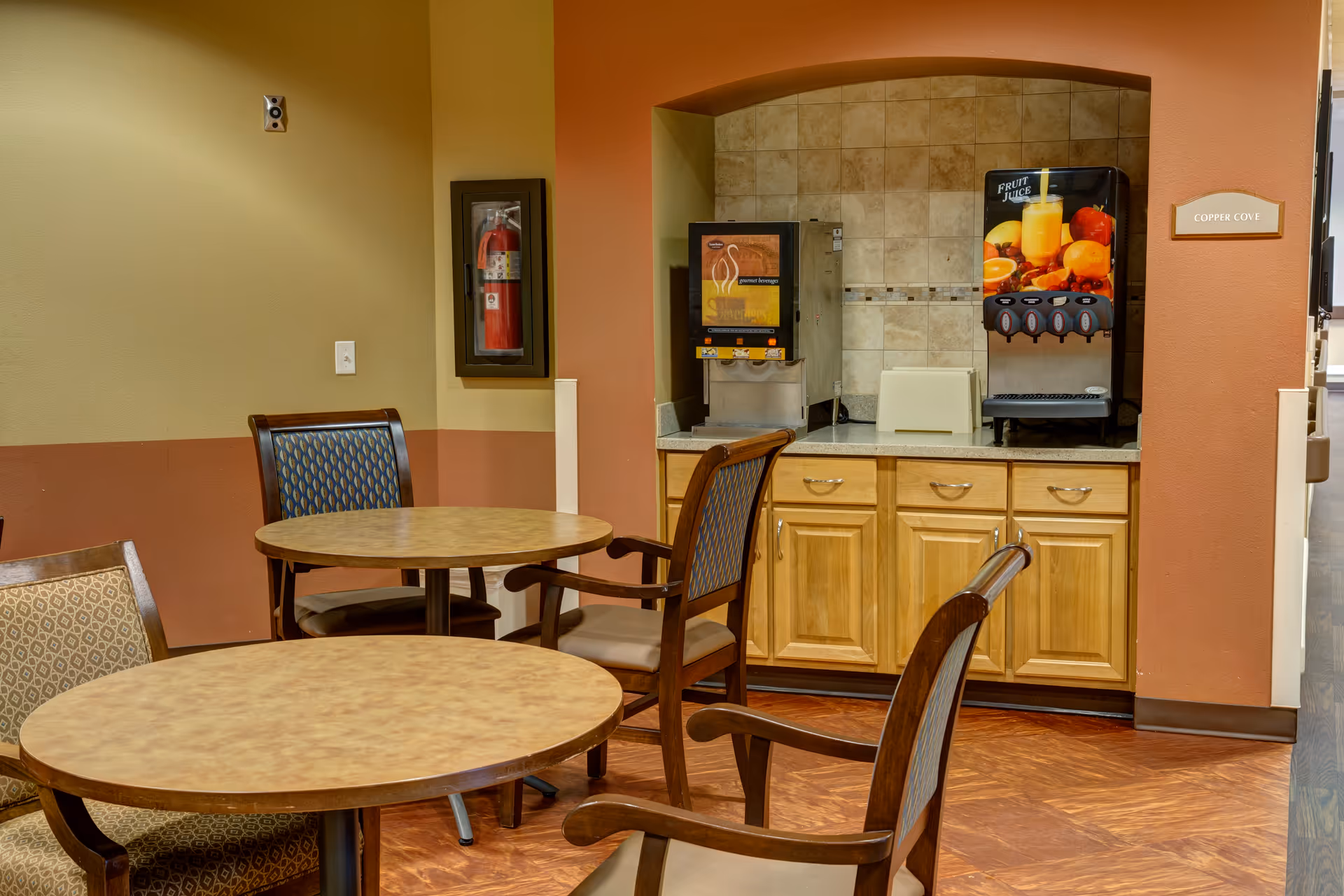 A small dining area with round tables and cushioned chairs. In the background, there is a countertop with a beverage dispenser and a juice machine. A fire extinguisher is mounted on the wall, and a sign labeled 'Copper Cove' is visible on the right side.