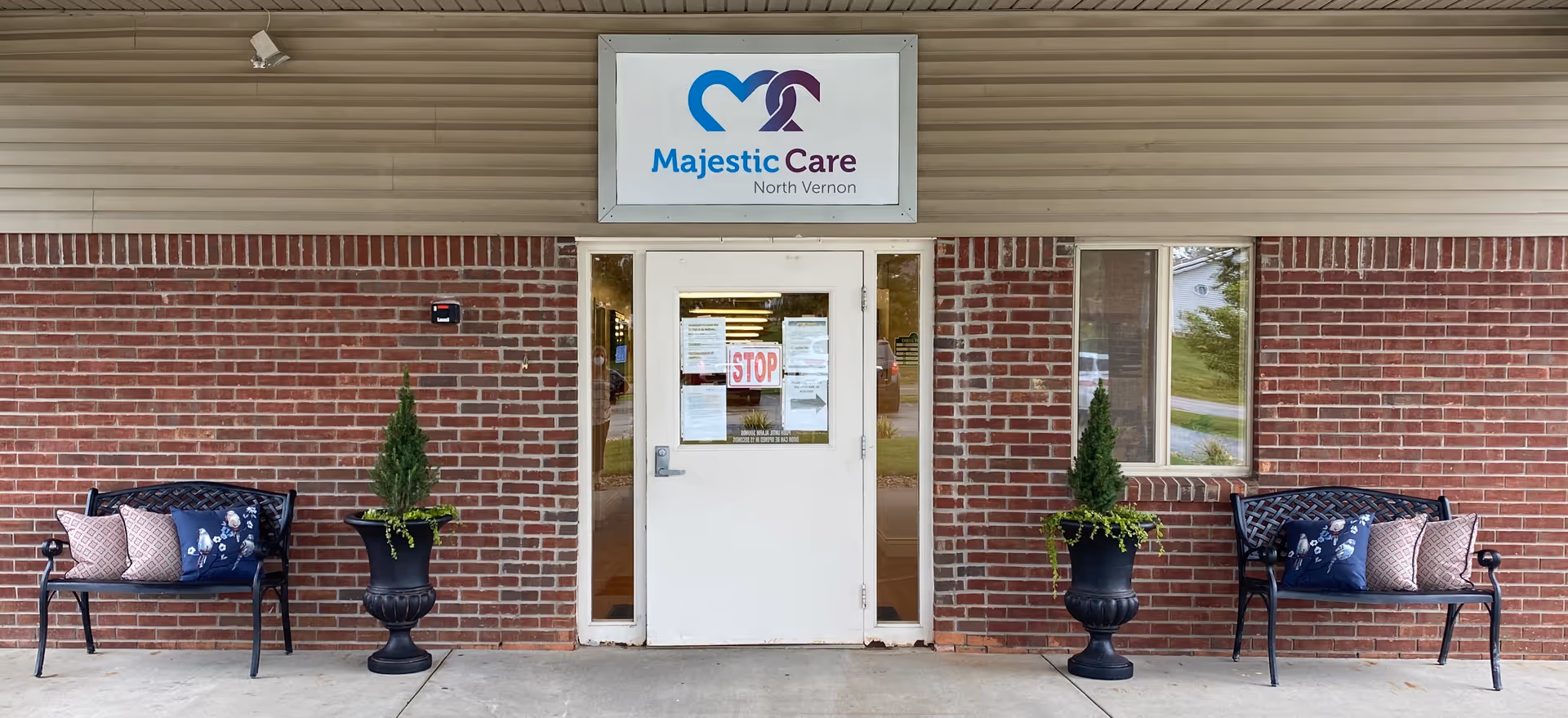 Entrance of Majestic Care of North Vernon facility with a white door centered between two windows. Above the door is a sign with the facility's logo and name. On either side of the door are black metal benches with decorative pillows and large black planters containing small evergreen trees and greenery. The building exterior is red brick with beige siding above.