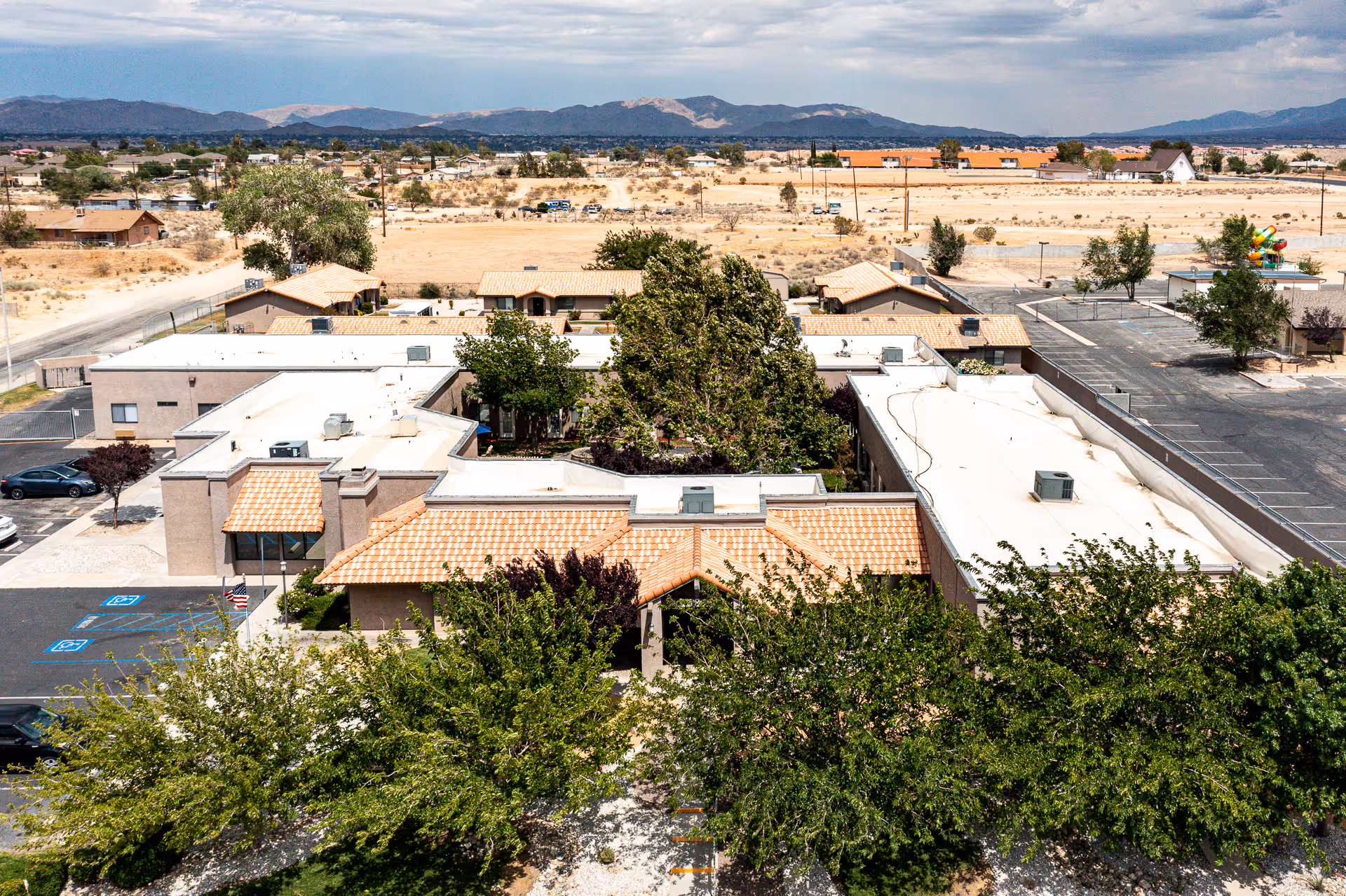 Aerial view of Sierra Vista Independent & Assisted Living facility showing a single-story building with a tiled roof entrance surrounded by trees and parking spaces. The background includes a dry, desert-like landscape with mountains in the distance under a partly cloudy sky.