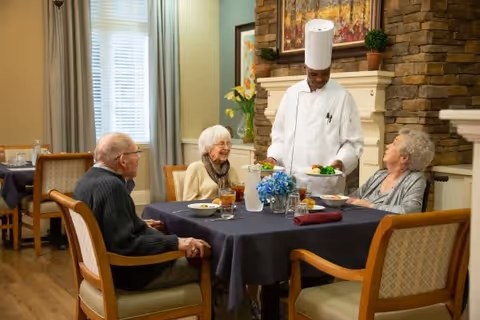 A chef in a white uniform and hat serving food to three elderly people seated around a dining table in a cozy room with a stone fireplace and framed artwork on the wall.
