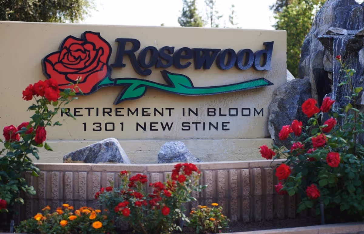 Entrance sign for Rosewood retirement community with a rose logo and red flowers in front.