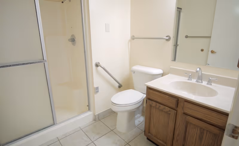 Bathroom in a senior living facility with a shower, toilet, sink, and wooden vanity.