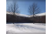 Snow covers the ground in front of a brick building with leafless trees.