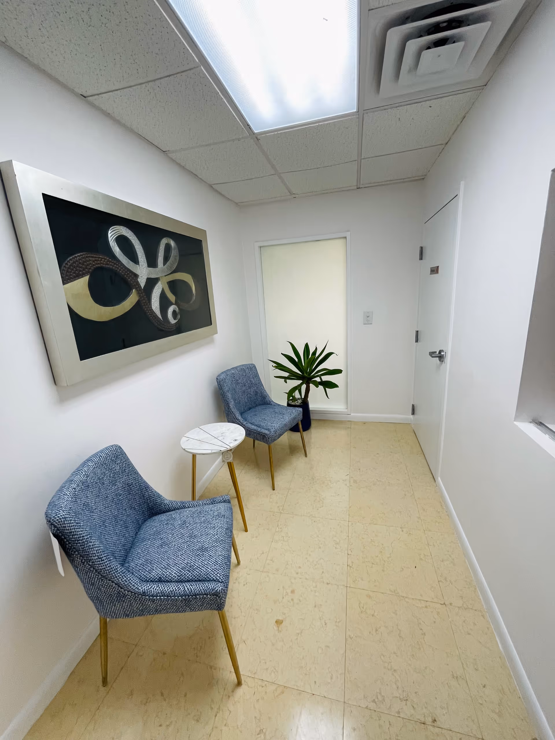 Small waiting area with two blue chairs, a round table, plant, and wall art.