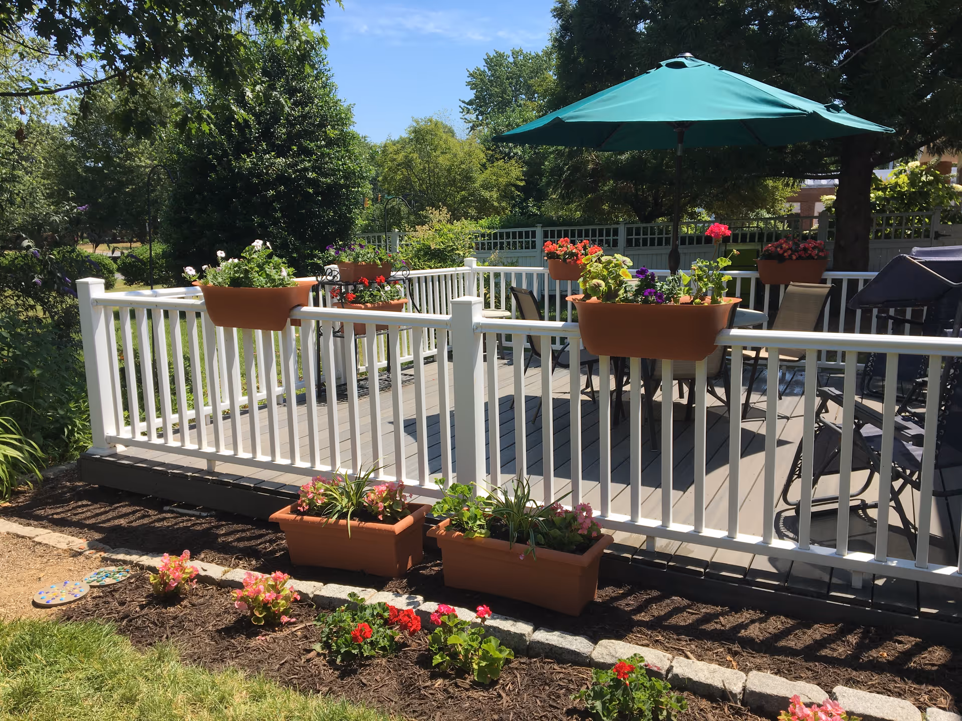Sunlit outdoor deck with white railing, potted flowers, a table with a green umbrella, and surrounding garden beds.
