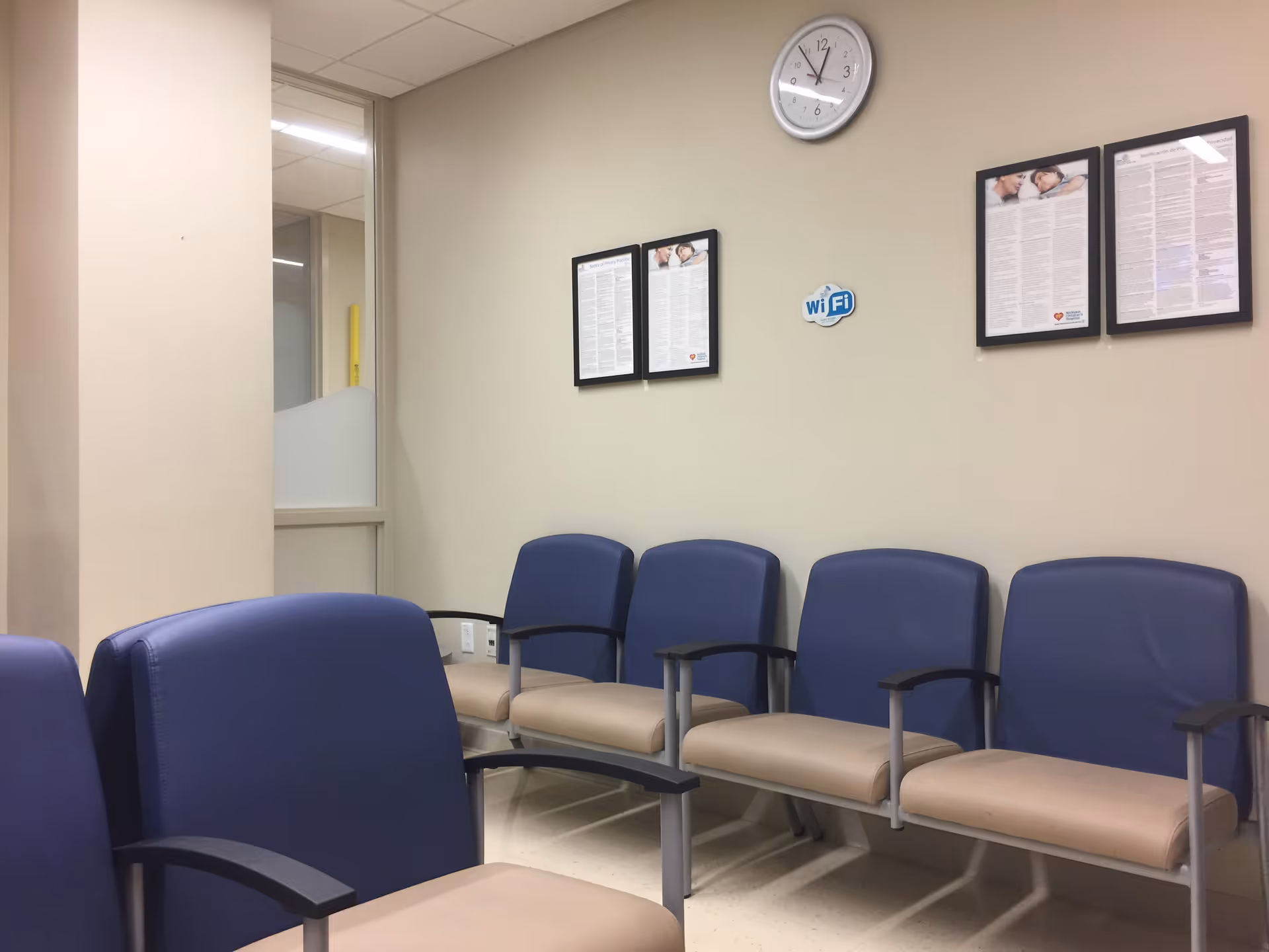Clinic waiting area with blue chairs, wall clock, and framed notices.