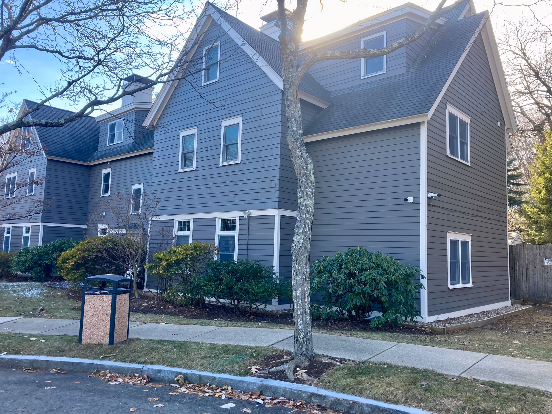 Gray multi-story house-like assisted living building with white-trimmed windows, a tree, shrubs, sidewalk and a trash bin in front.