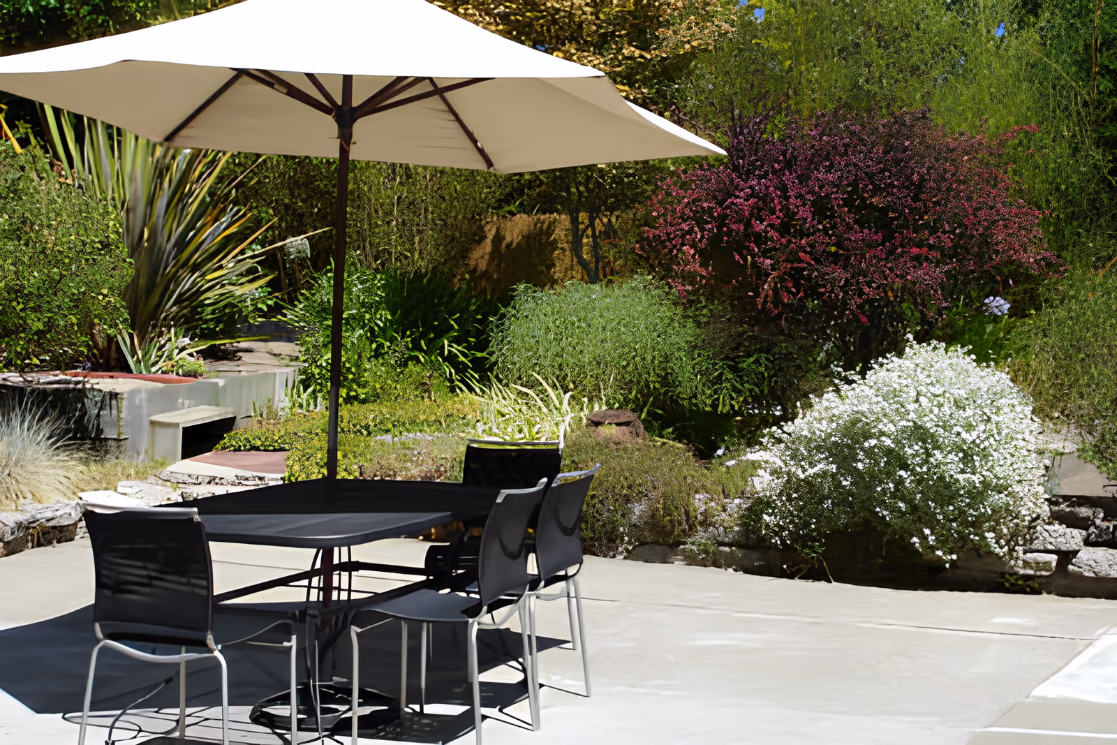 Outdoor patio area with a table and four chairs under a large white umbrella, surrounded by lush greenery and flowering bushes.