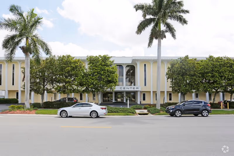 Exterior view of a beige senior living center with palm trees and parked cars.
