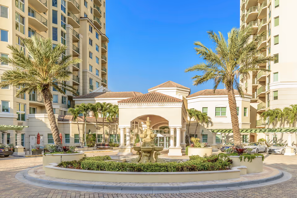 Exterior view of Vi at Aventura senior living facility showing a large entrance with a tiled roof, a central fountain surrounded by greenery, palm trees, and tall residential buildings on either side under a clear blue sky.