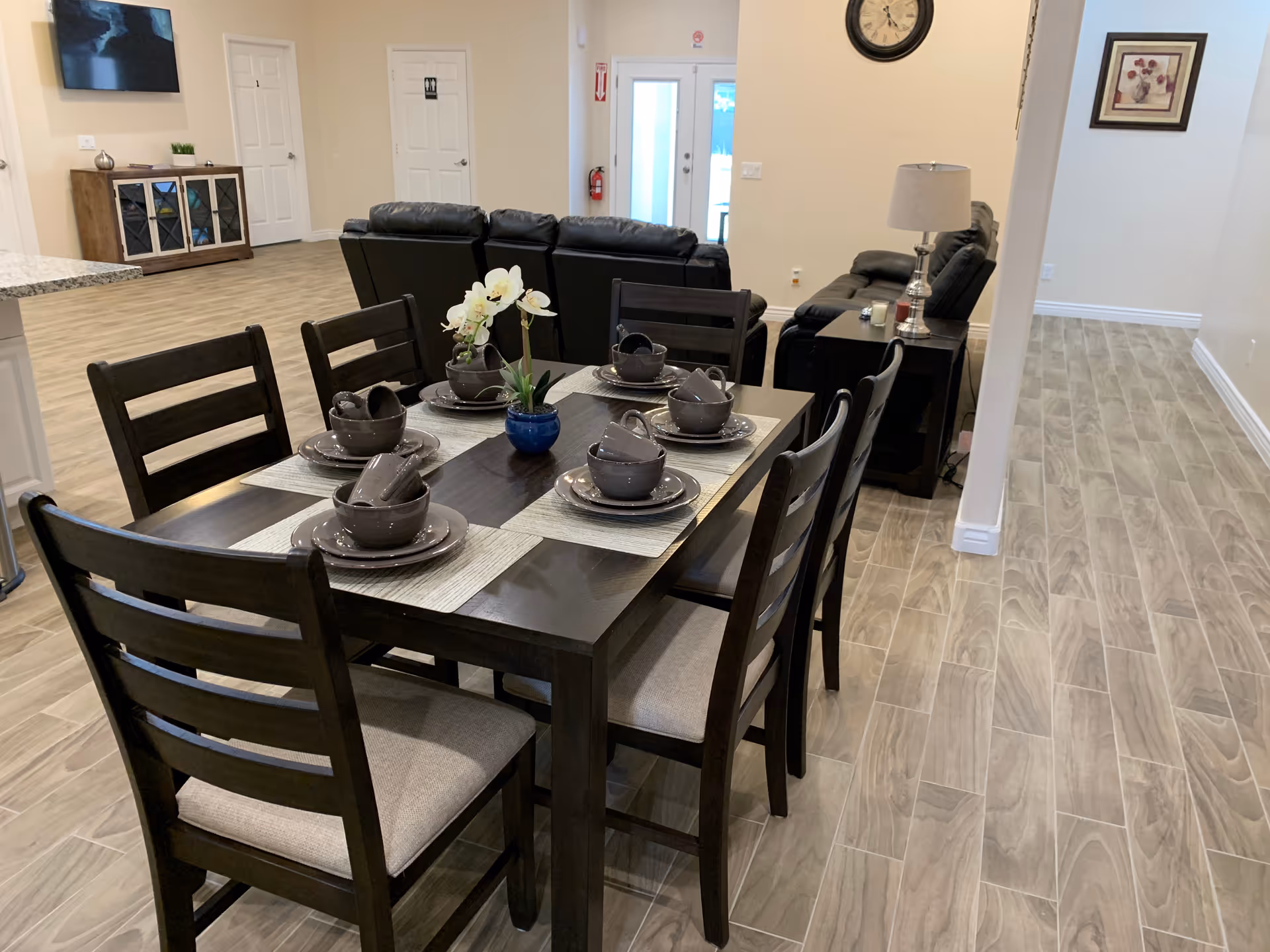 A dining area in an assisted living facility with a dark wooden table set for six people, each place setting including a plate, bowl, and cup. A small potted orchid is in the center of the table. Behind the dining area is a living room with black leather sofas, a side table with a lamp, a wall clock, and a framed picture. The floor is covered with wood-patterned tiles, and there is a TV mounted on the wall above a cabinet in the background.