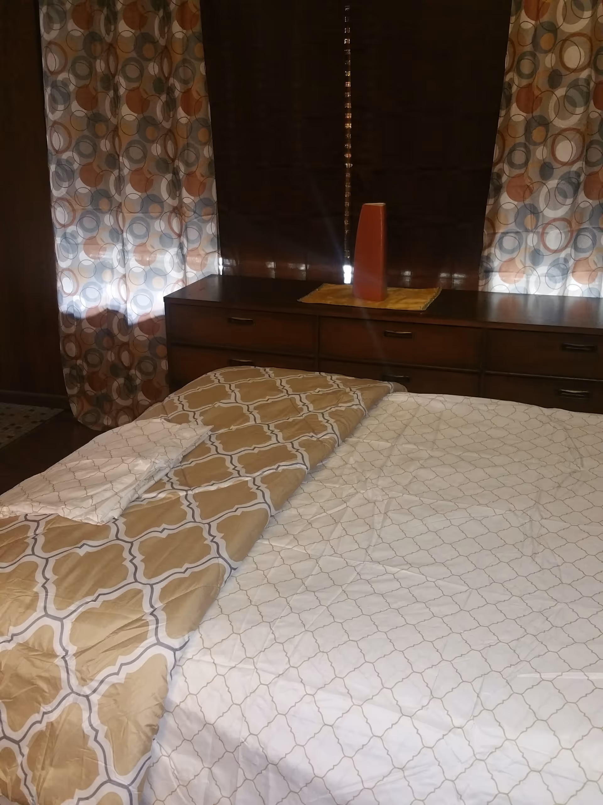 A neatly made bedroom with a patterned bedspread and wooden dresser.