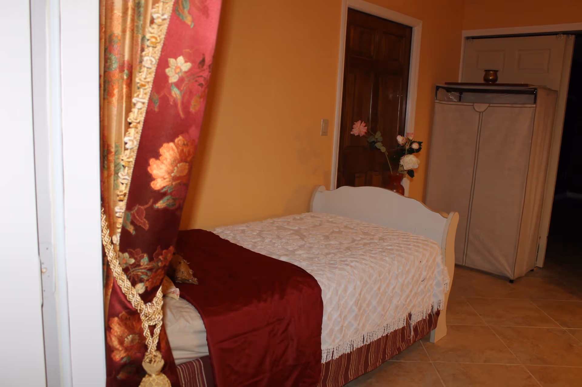 A small bedroom with a single bed covered with a white textured blanket and a red throw. There is a floral curtain tied back on the left side, a wooden door in the background, a vase with artificial flowers on a small table, and a beige fabric wardrobe on wheels. The walls are painted yellow and the floor is tiled.