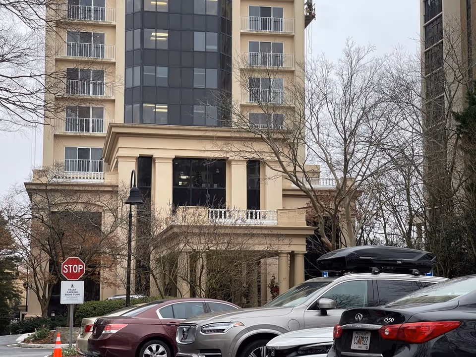 Front exterior of a multi-story beige residential building with parked cars, trees, and a stop sign.