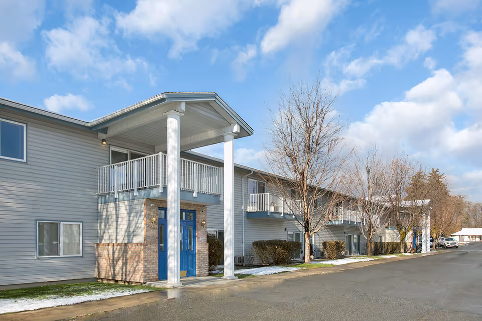 Exterior view of a two-story senior living facility building with light gray siding and blue doors. The building has balconies on the upper floor and leafless trees along the sidewalk. The sky is partly cloudy with patches of blue.
