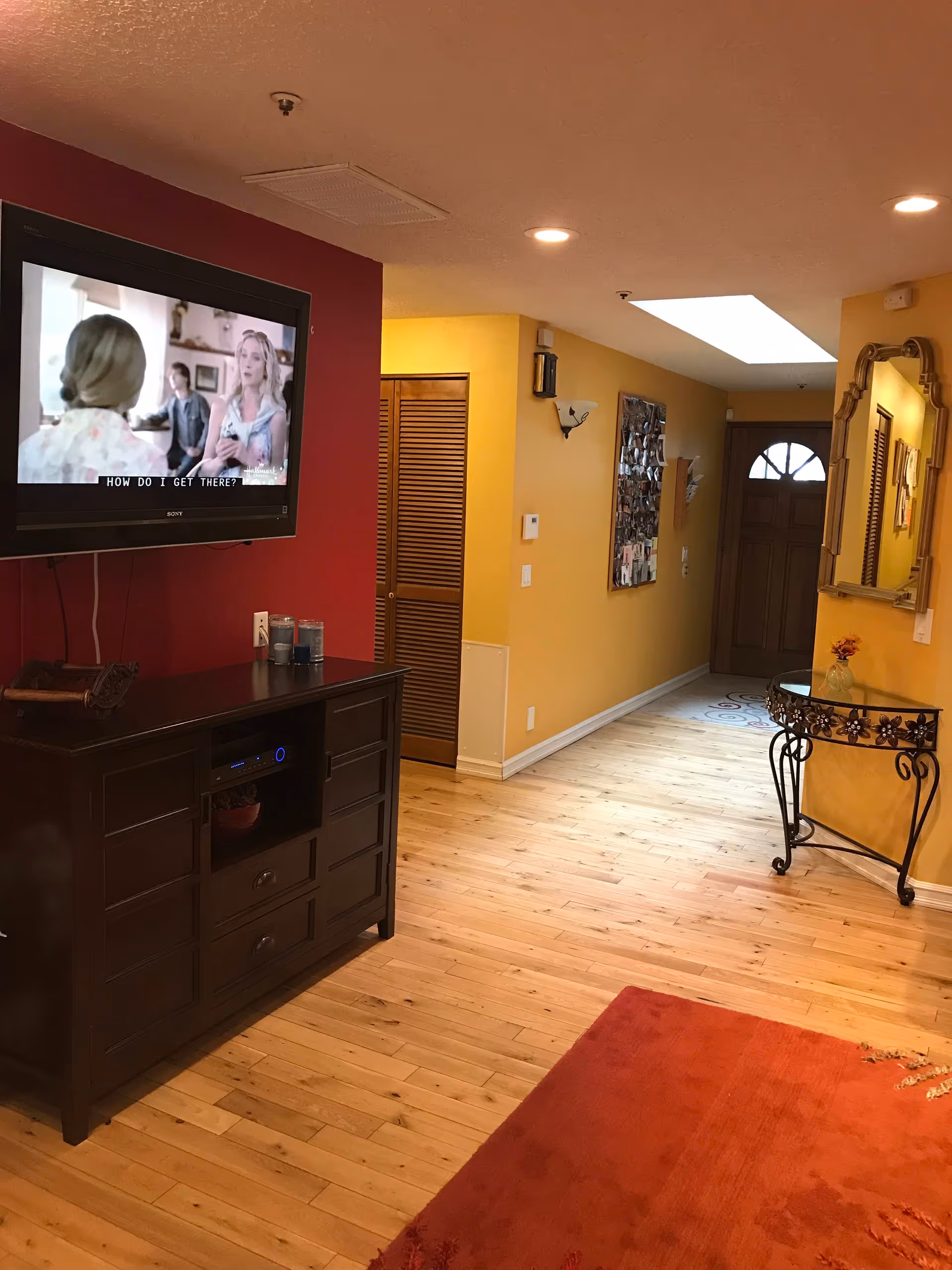 Interior view of a hallway in a senior living facility with yellow walls and wooden flooring. A flat-screen TV is mounted on a red accent wall above a dark wooden cabinet. The hallway leads to a wooden front door with a decorative glass window. A small glass table with a floral design and a vase with flowers is positioned against the right wall beneath a large ornate mirror. A bulletin board with photos is visible on the left wall of the hallway.