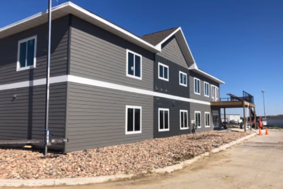 Exterior view of a two-story building with gray siding and white trim under a clear blue sky. The building has multiple windows and a small elevated wooden deck with stairs on the right side. The ground around the building is covered with rocks and there are orange traffic cones near the deck.