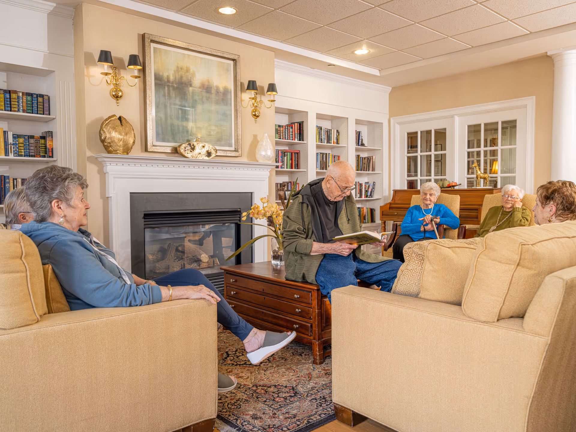 A group of elderly residents seated in a cozy assisted-living common room with a fireplace and bookshelves while a man reads from a book.