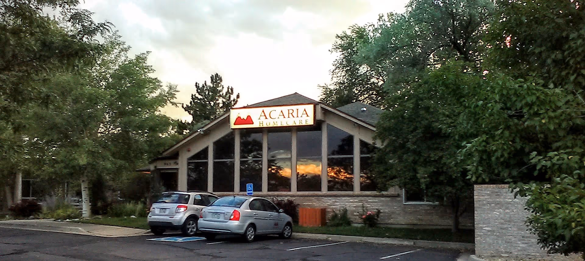 Acaria Homecare building front with large windows, sign, and parked cars in parking lot.