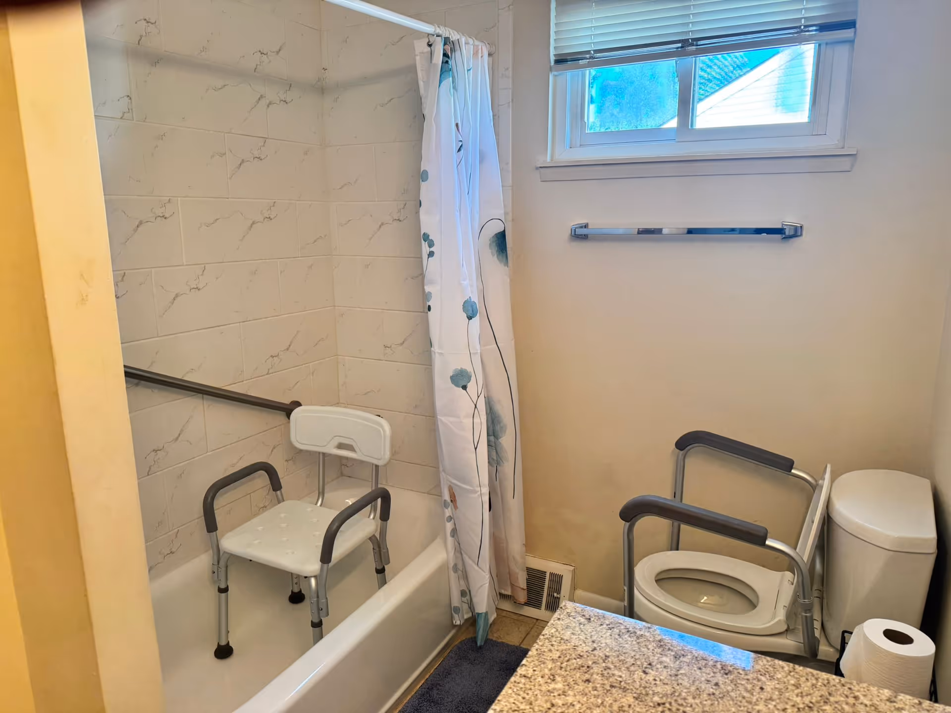 Accessible bathroom with tub, shower chair, grab bars, toilet, and a window.