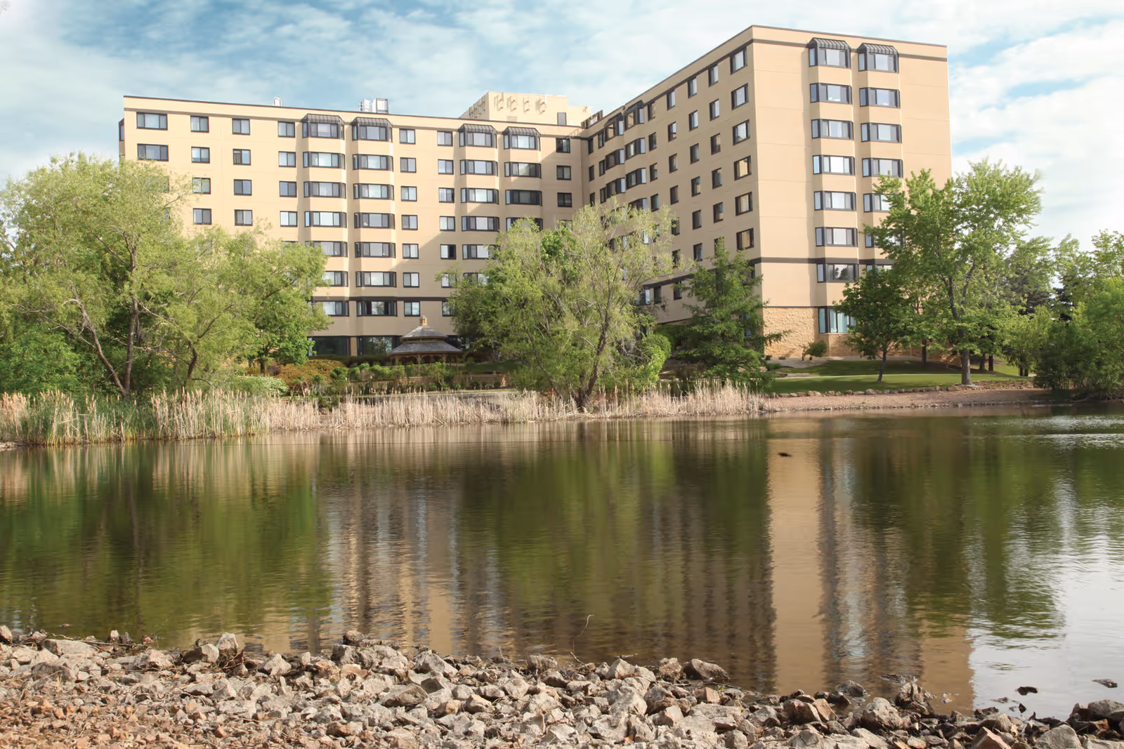 Exterior view of a beige multi-story senior living building beside a lake with trees.