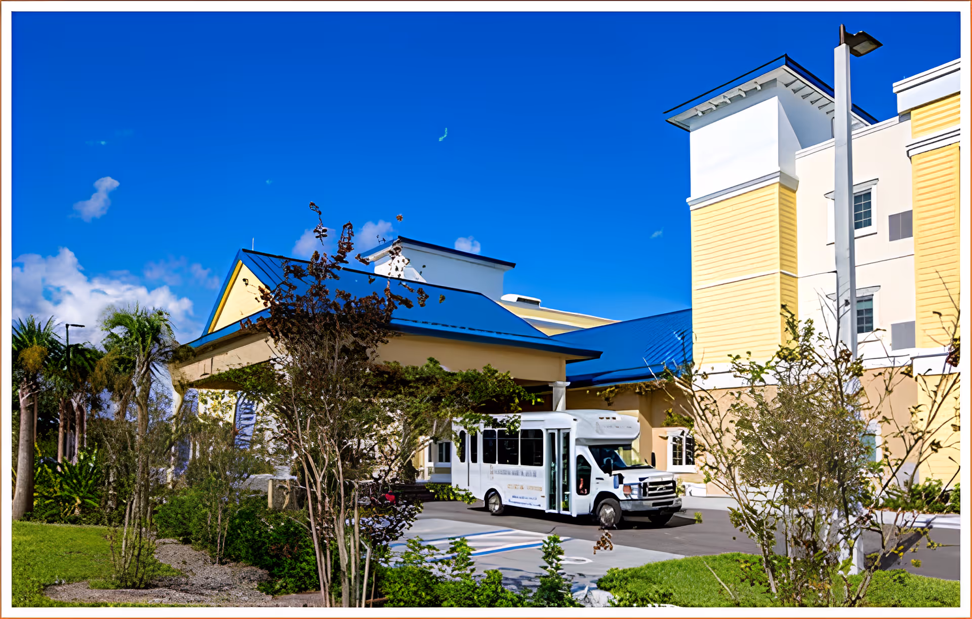 Front entrance of a yellow multi-story senior living building with blue roofs, landscaping, and a white shuttle bus parked under the porte-cochere.
