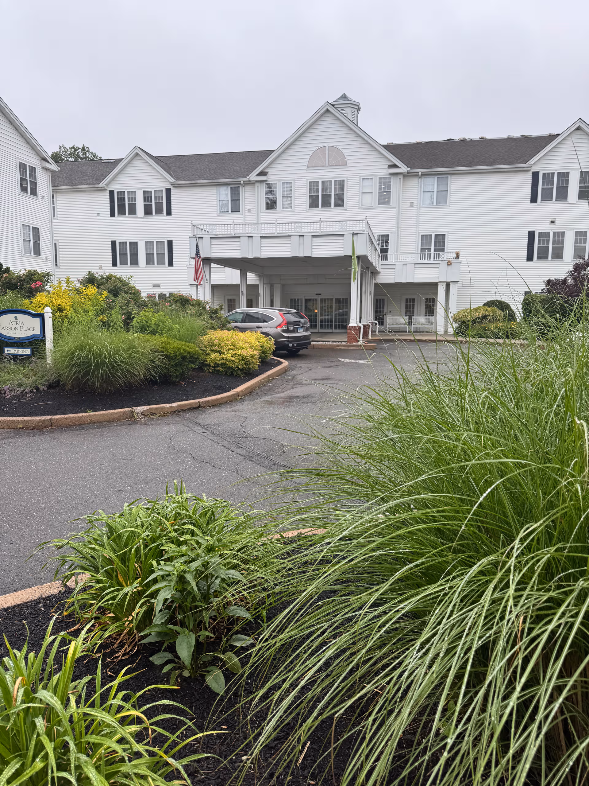 White, multi-story senior living building with a car parked near the entrance and landscaping.