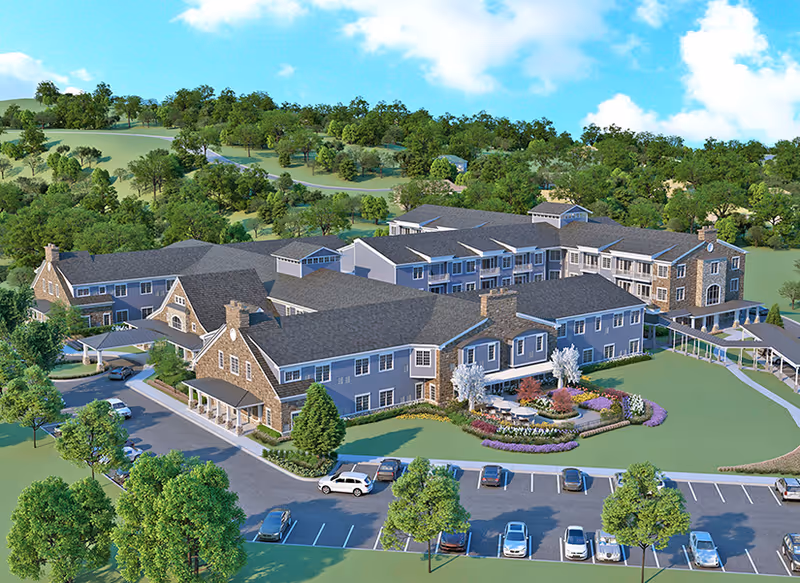 Aerial view of Artisan at Hudson, a large senior living facility with multiple connected buildings featuring gray roofs and stone accents. The facility is surrounded by landscaped gardens, trees, and parking areas with several cars. The background shows rolling green hills under a blue sky with scattered clouds.