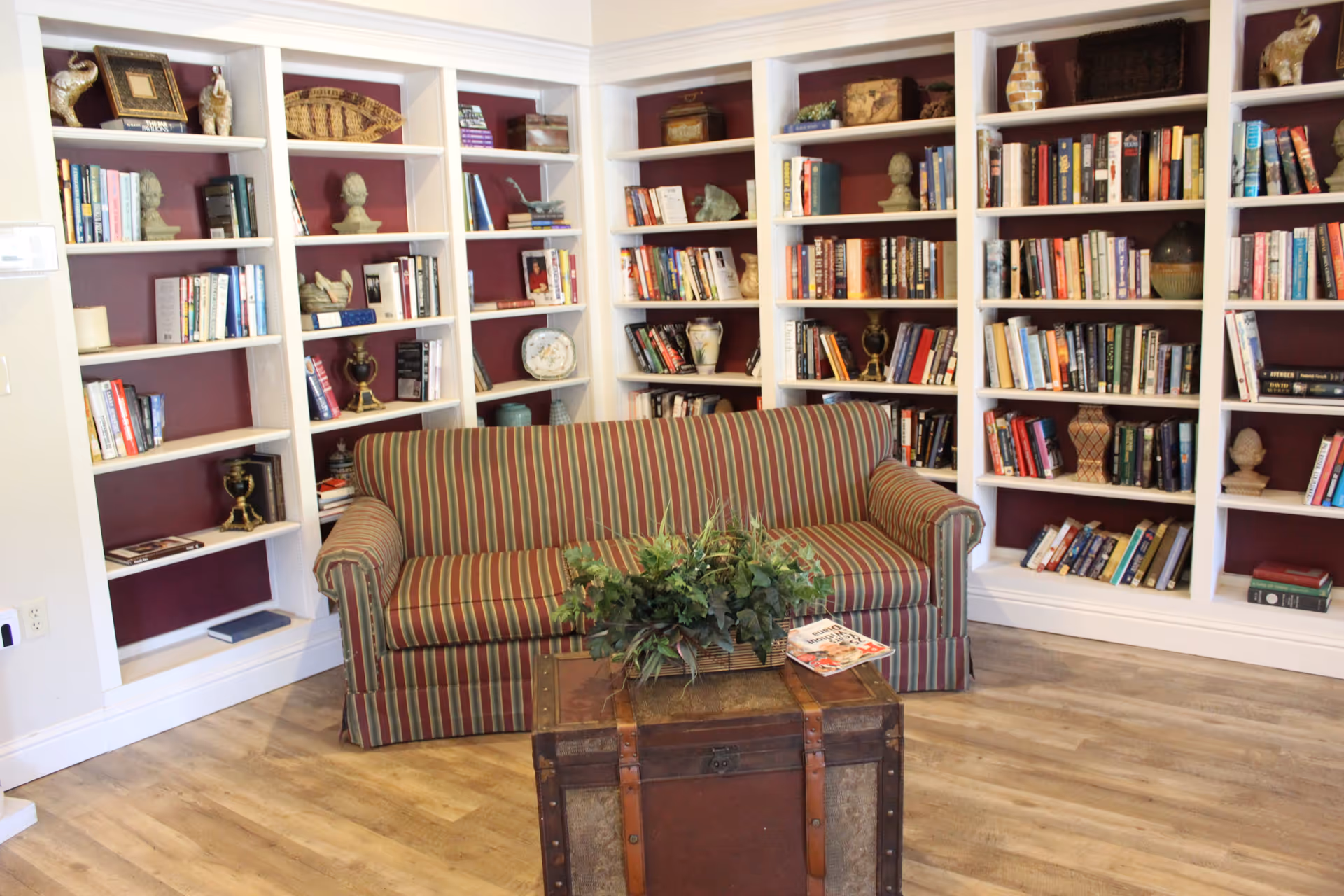 A cozy reading nook with a striped sofa in front of built-in white bookshelves filled with books and decorative items. A wooden chest serves as a coffee table with a plant and magazines on top. The floor is light wood, and the walls behind the shelves are painted a deep red.