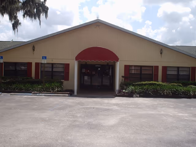 Front view of a single-story senior living center with a red awning and shrubs.