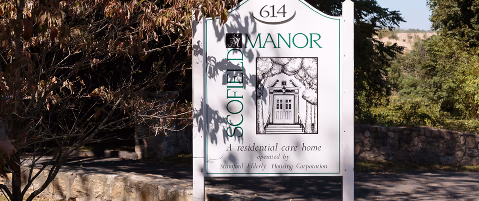 White sign for Scofield Manor residential care home (614) displayed outdoors near trees and a stone wall.