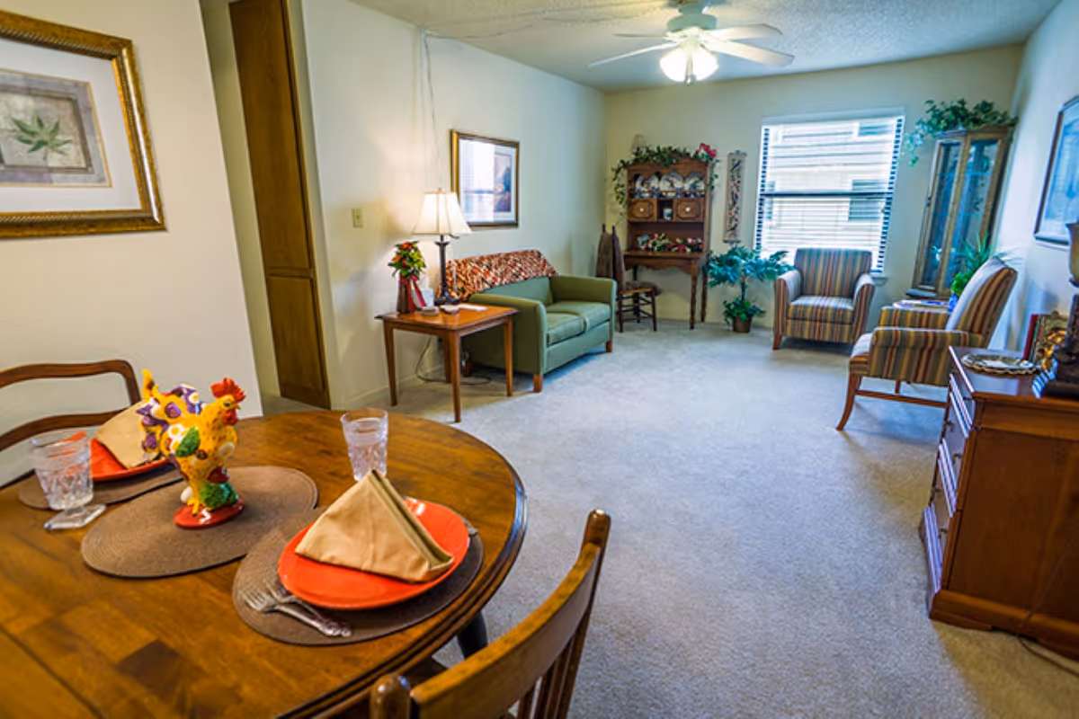 Bright senior living common area with round dining table, seating, and plants.